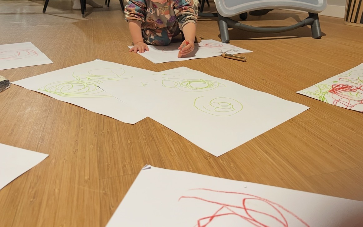 Young toddler sitting on floor surrounded by papers with green circular scribbles showing early mark-making and motor skill development